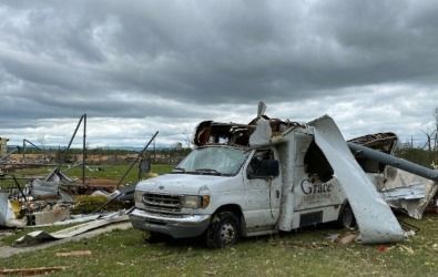 Damaged white van with torn roof and sides; debris-filled landscape, cloudy sky, and buildings.