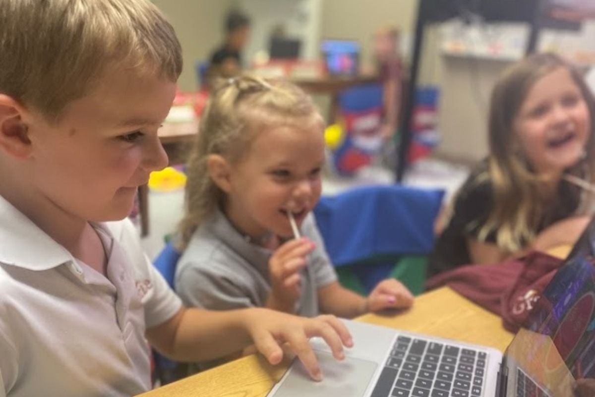 Three children at a table using a laptop, one with a lollipop, smiling. Classroom setting.