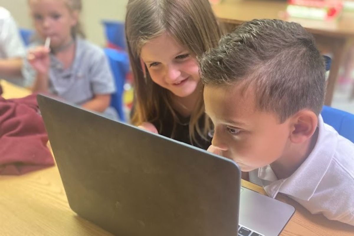 Two children, a girl and a boy, looking at a laptop screen, girl smiling, in a classroom setting.