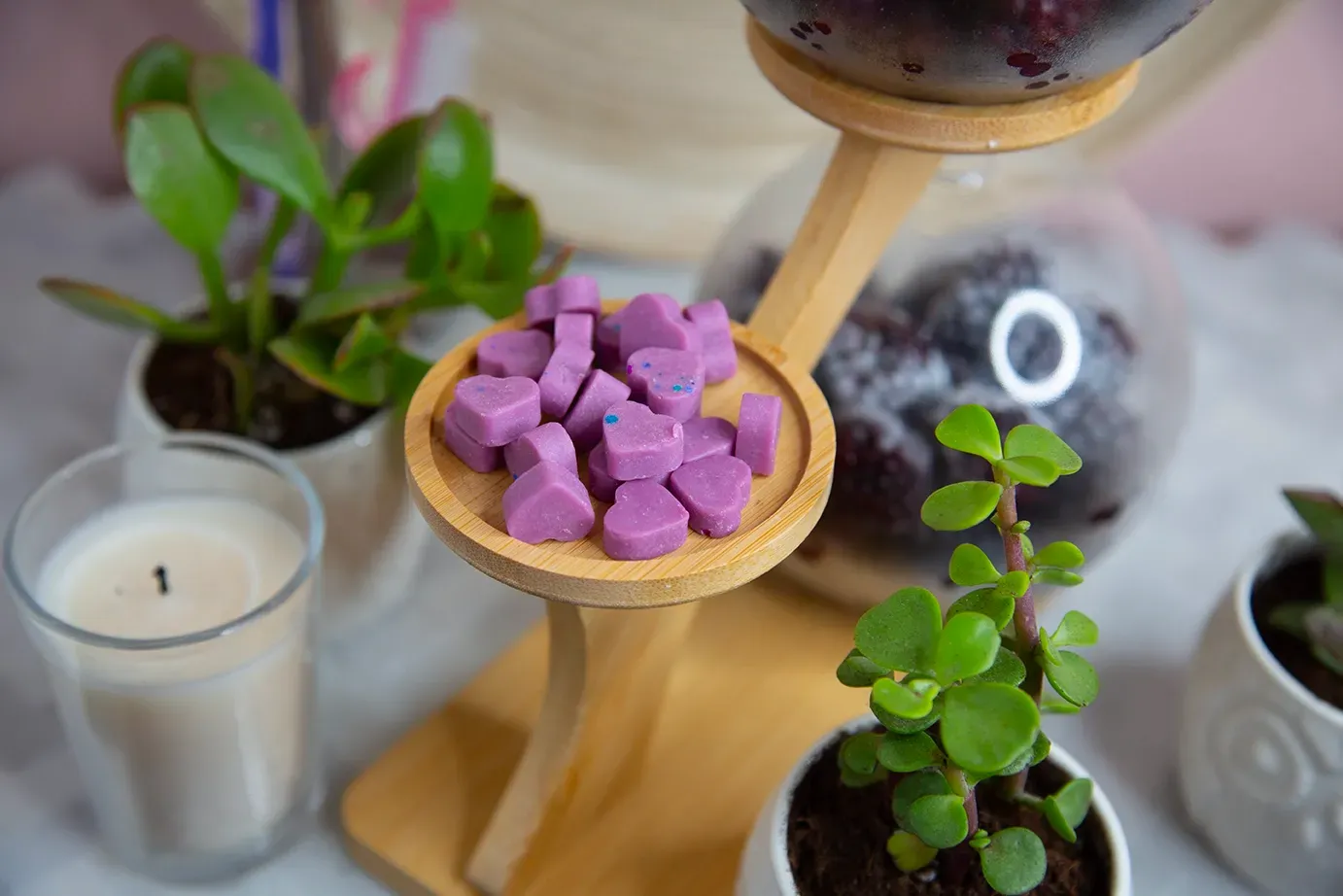 Purple wax melts are sitting on a wooden tray on a table next to potted plants and candles.