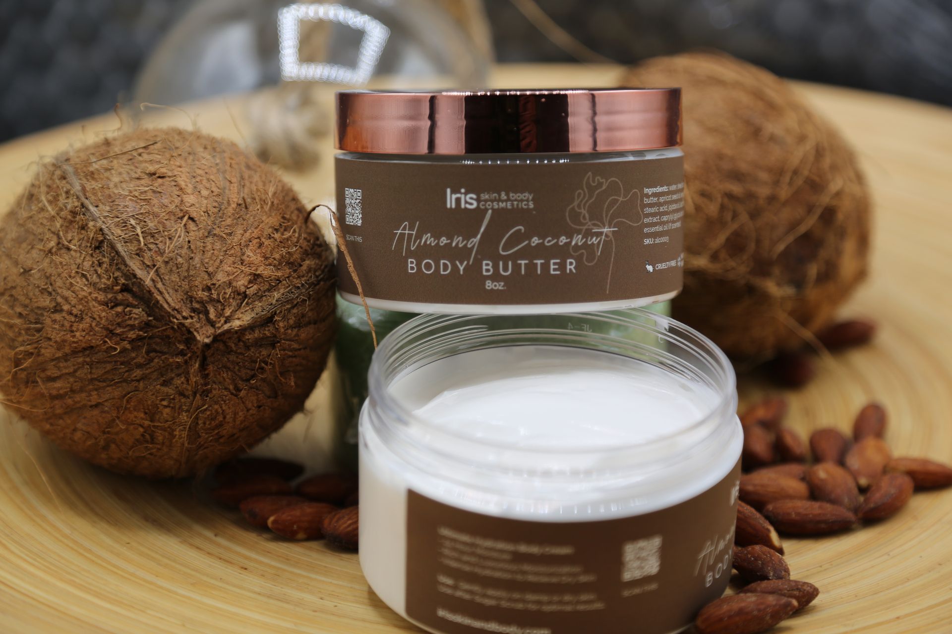 A jar of body butter is sitting on a wooden table next to coconuts and almonds.