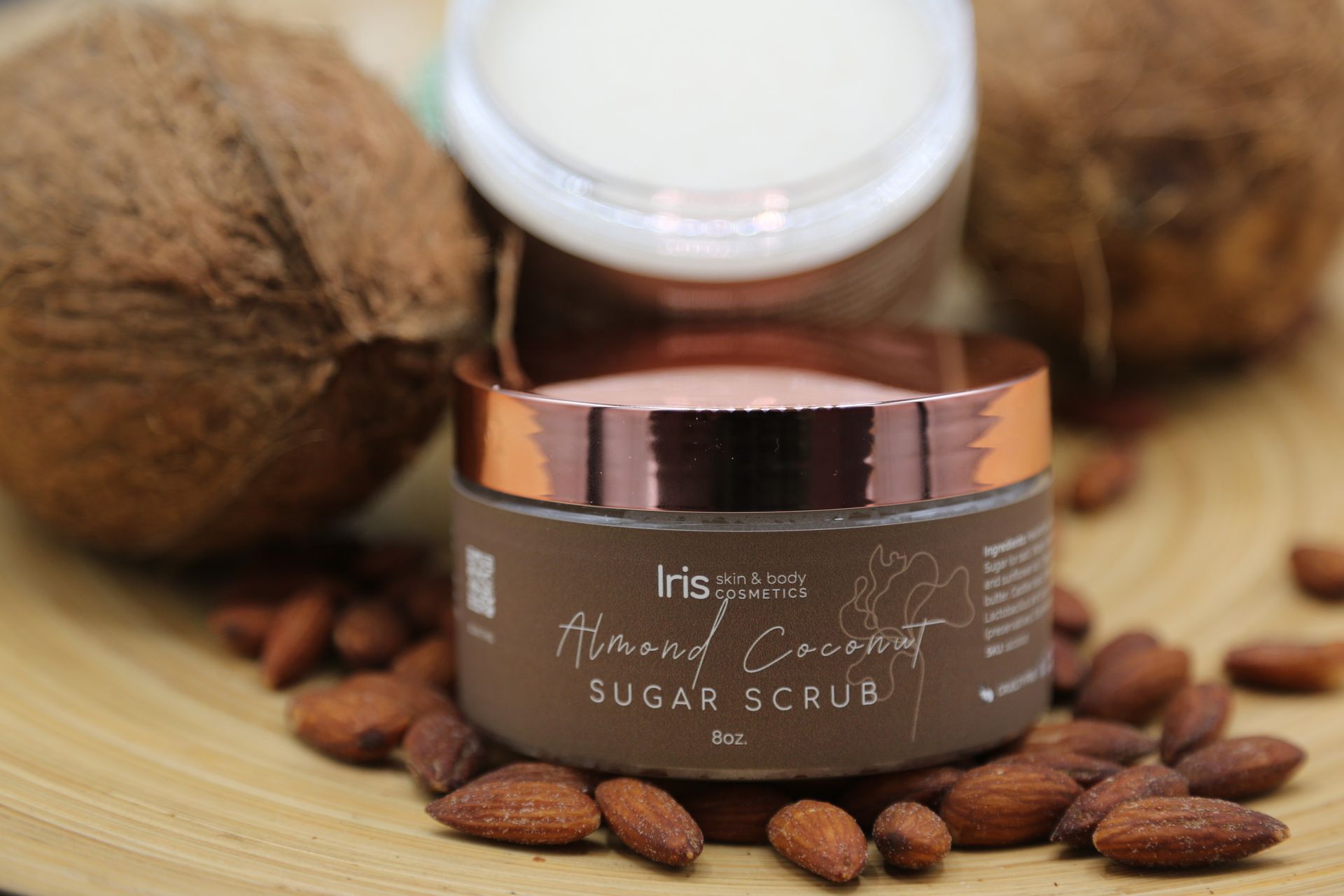A jar of sugar scrub is surrounded by almonds and coconuts.