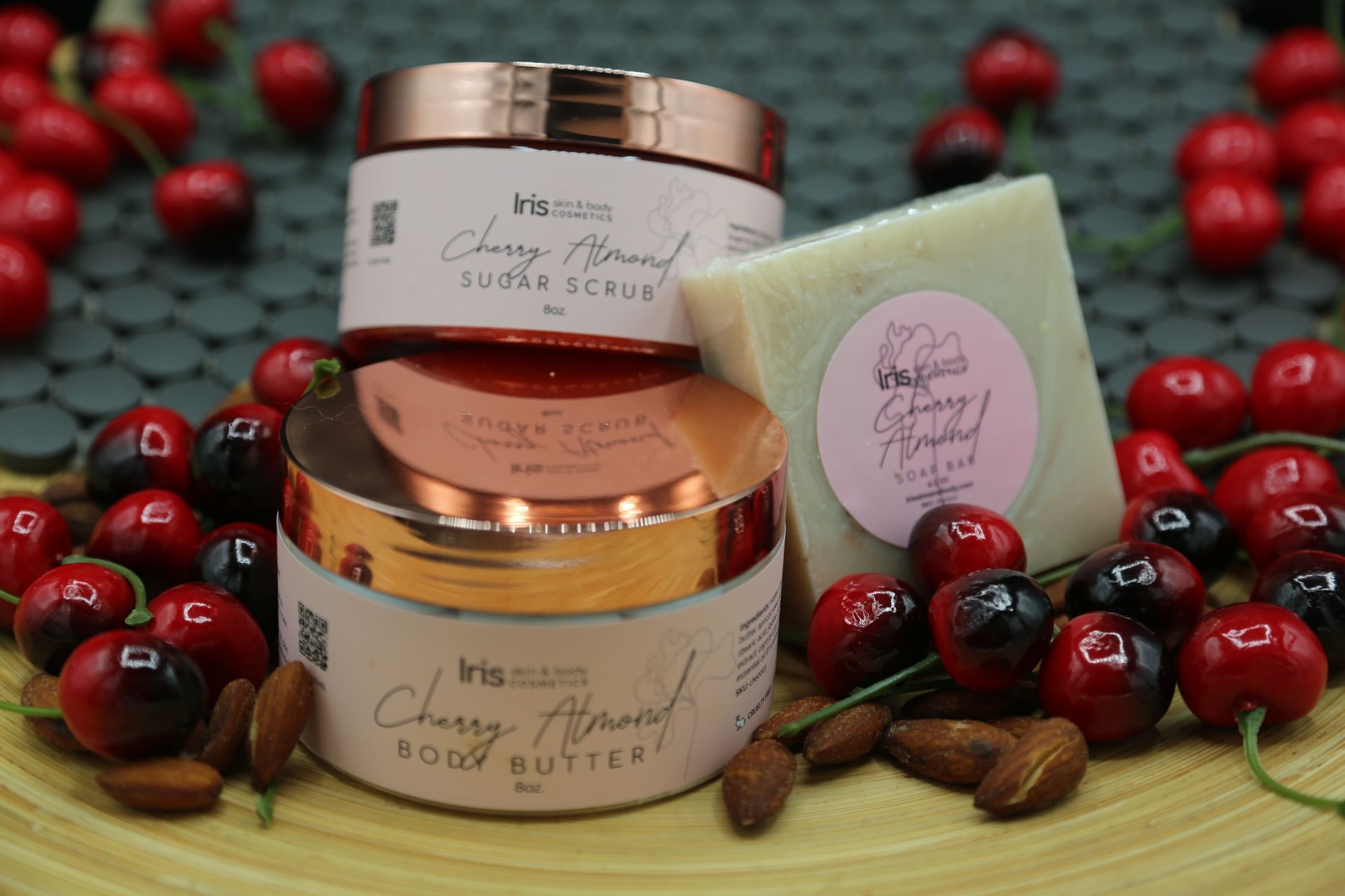 A cherry almond body butter , sugar scrub , and soap are sitting on a table surrounded by cherries.