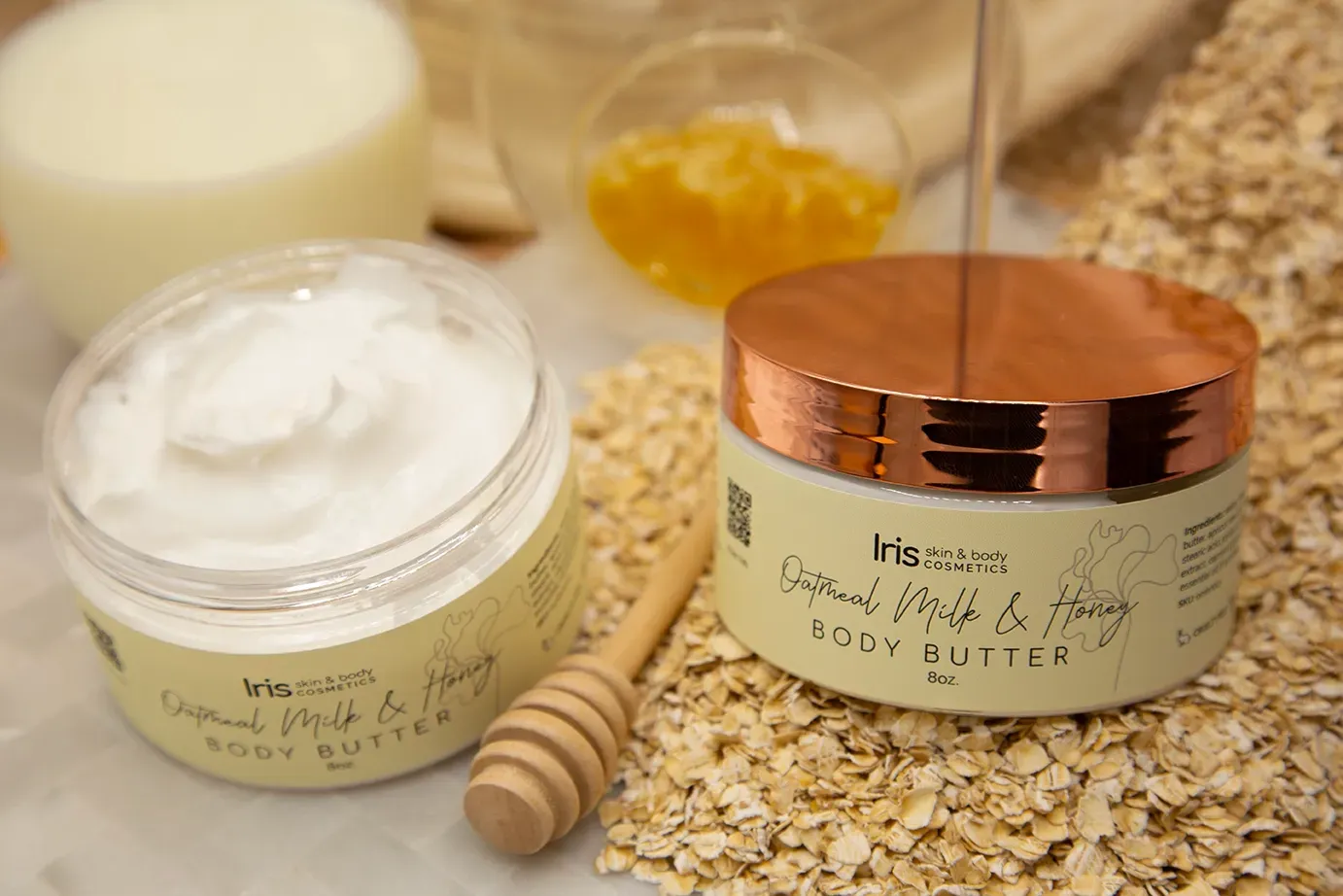Two jars of body butter are sitting on a table next to oats and honey.