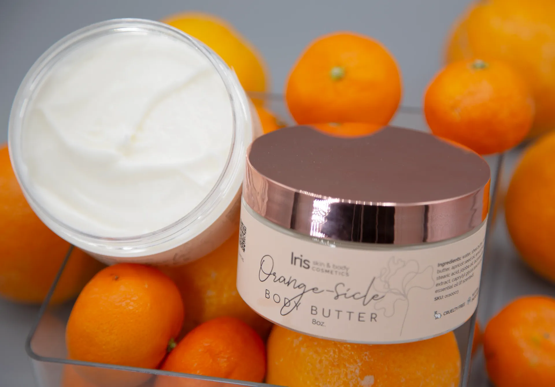 A jar of orange scented body butter is surrounded by oranges