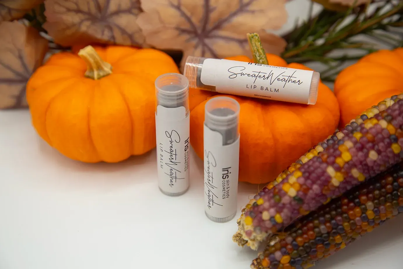 Two tubes of lip balm are sitting next to pumpkins and corn on the cob.