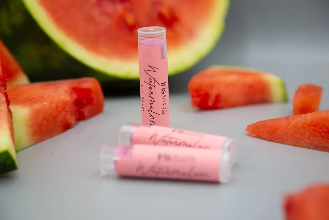 Three watermelon lip balms are sitting on a table next to watermelon slices.