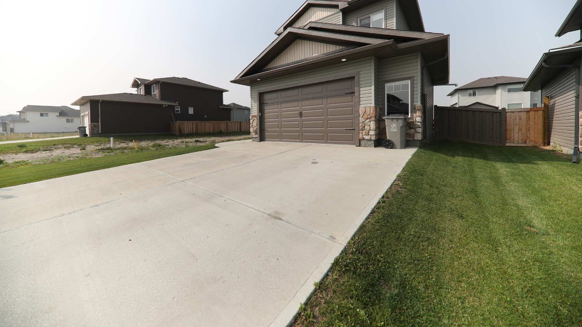 A beige multi-story house with a two-car garage, concrete driveway, and green lawn in a suburban neighborhood.