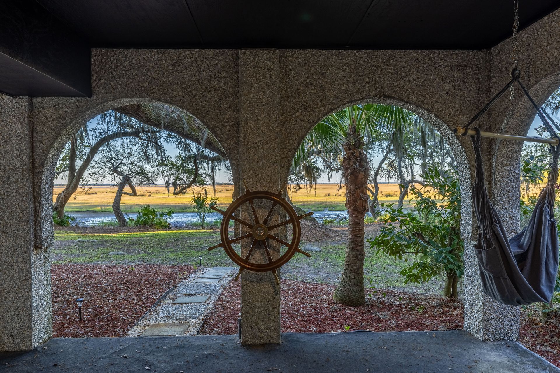 Arched porch with a ship wheel and hammock overlooking a grassy area, trees, and water.
