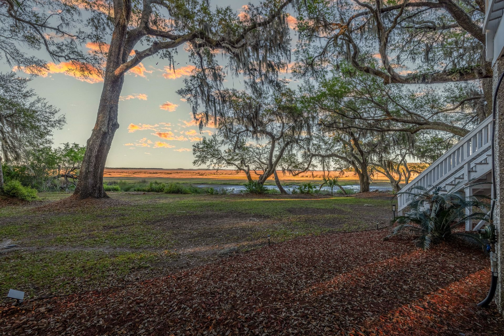 Grassy yard with trees, moss, and a glimpse of water at sunset; white porch on right.