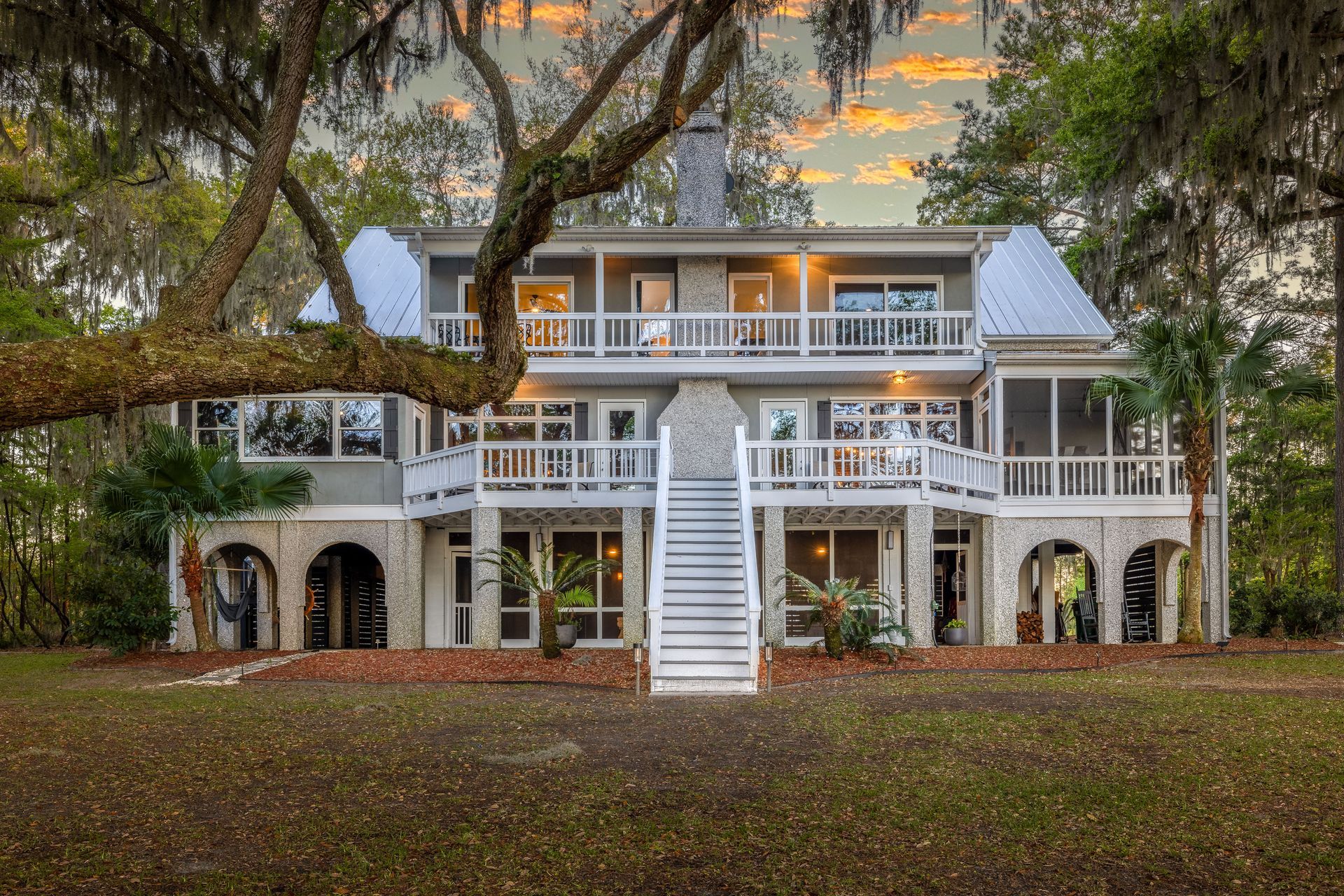 Three-story waterfront house with white balconies and stairs, surrounded by trees and a sunset sky.