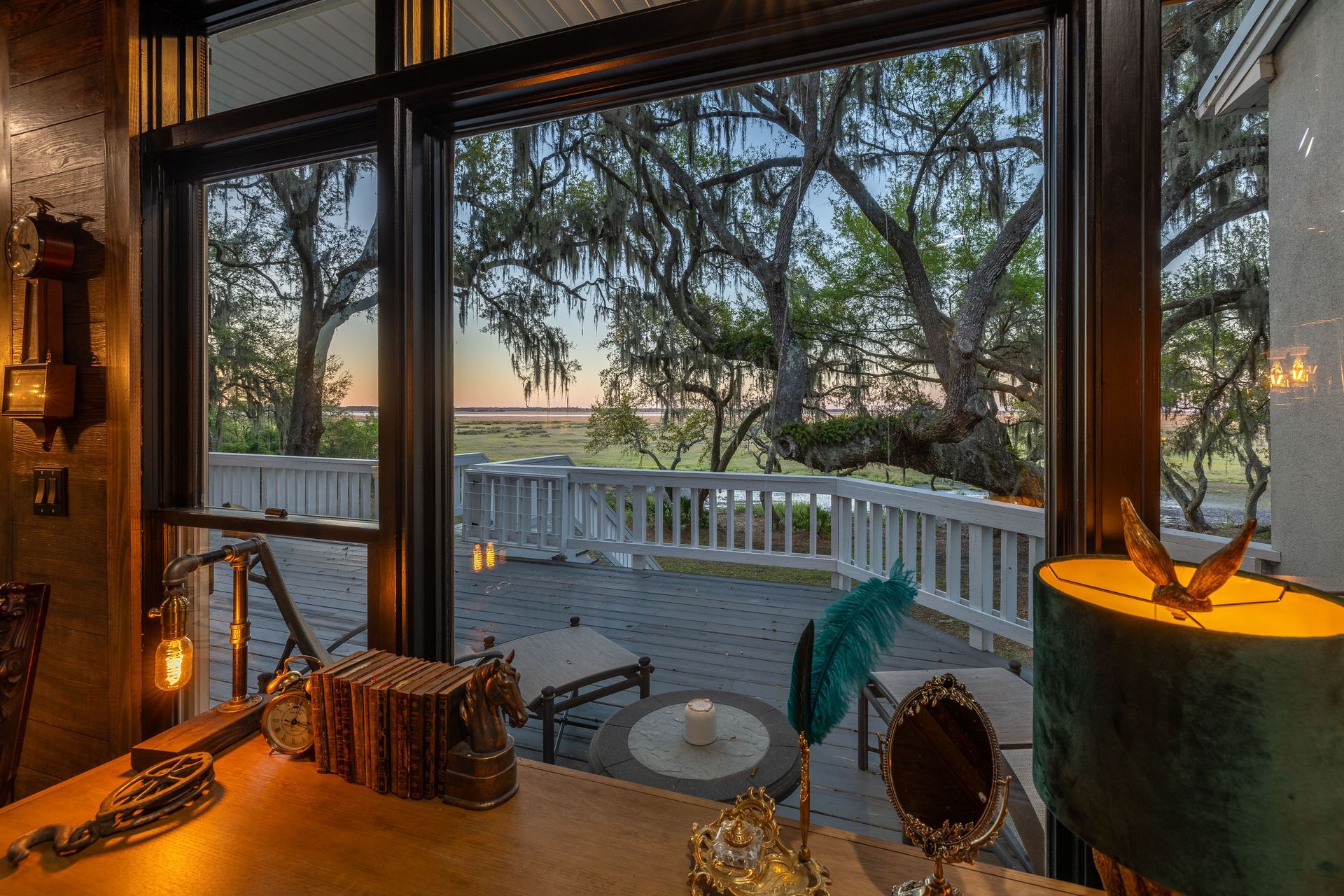 Interior with large windows overlooking a patio and trees at dusk. Desk with antique items is in the foreground.