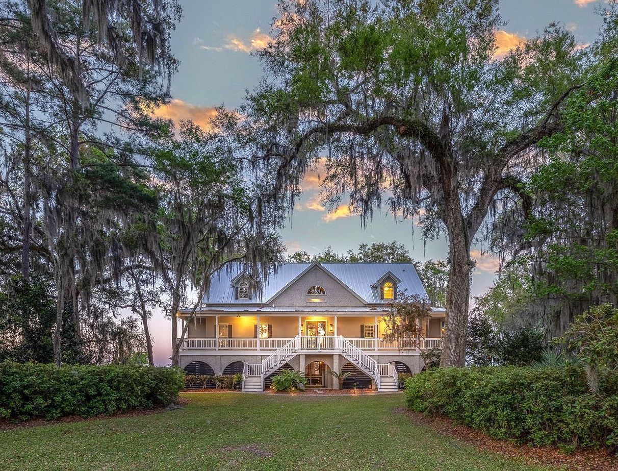 Elegant home framed by trees, with a porch, front steps, and a grassy lawn under a dusky sky.