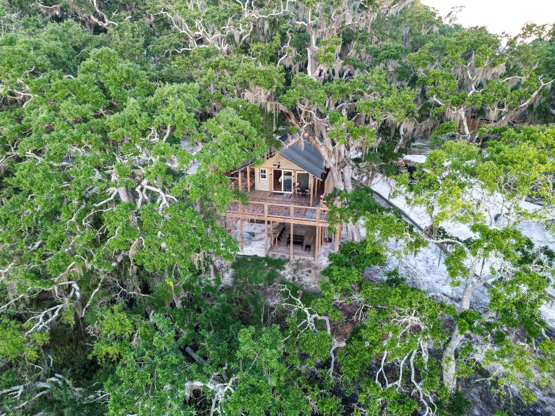 A wooden cabin with a deck nestled in lush green trees, overlooking a sandy area.