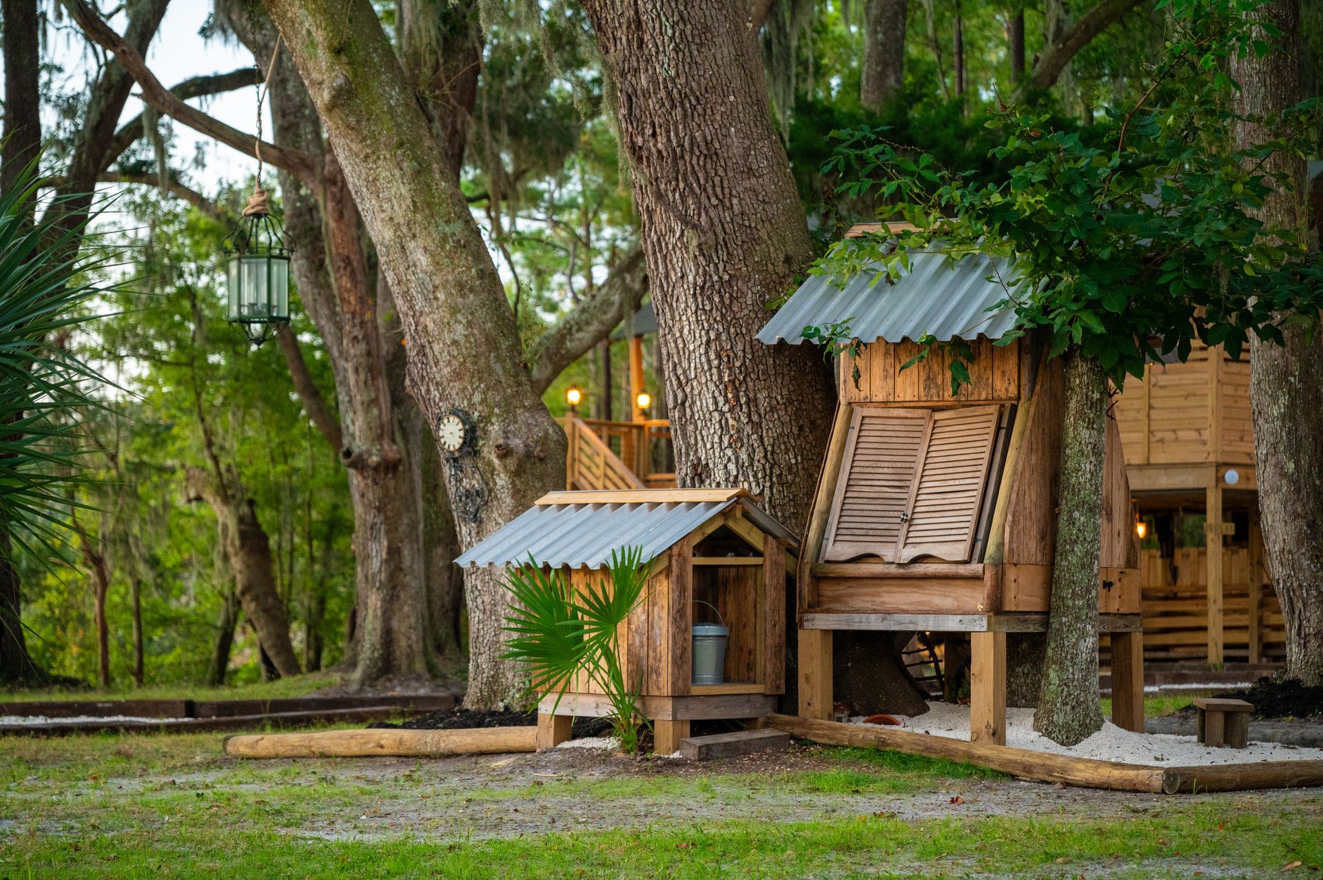Wooden treehouse structure in a grassy area, built around large trees with metal roofs.