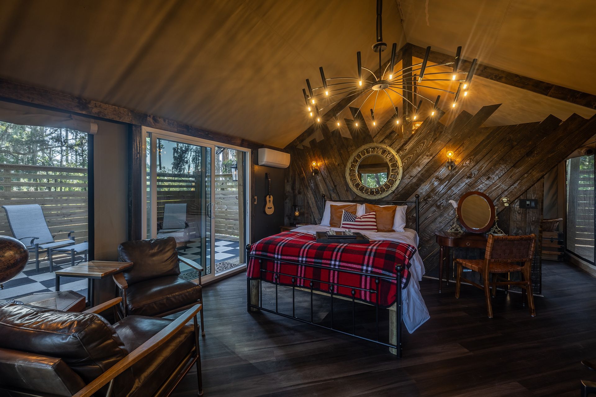Upscale glamping tent interior: bedroom with bed, seating, vanity, and outdoor patio access. Dark wood and modern lighting.