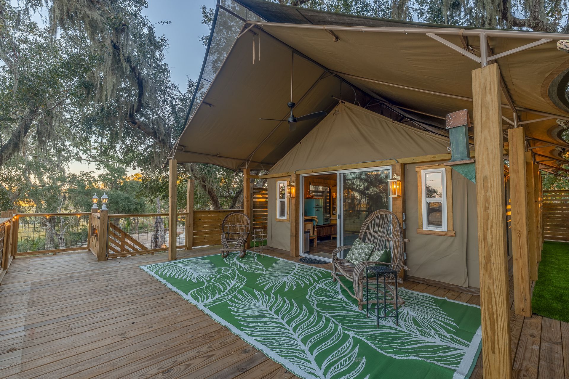 Glamping tent with a wooden deck, green rug, and two chairs. Exterior view in a wooded setting.