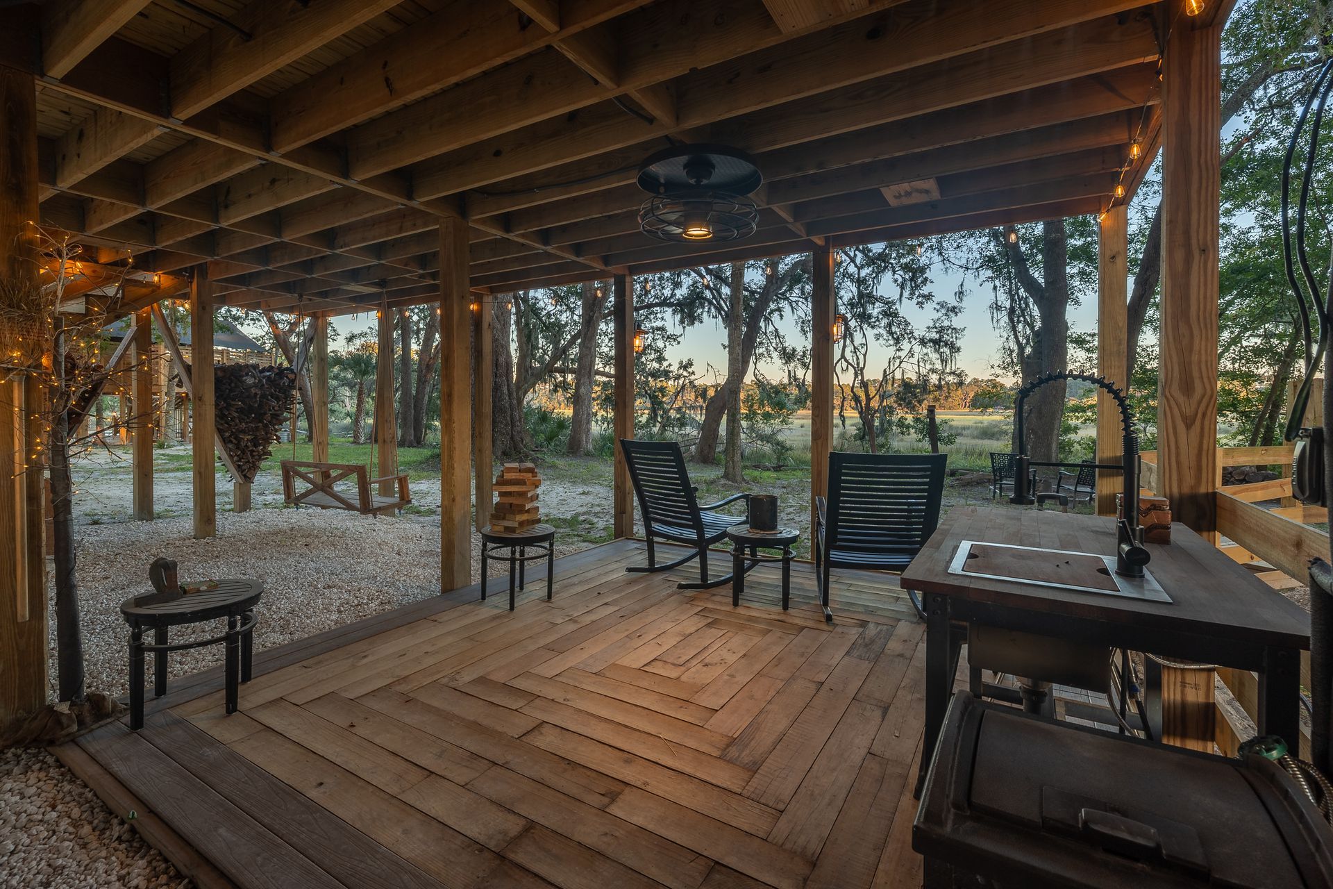 Covered outdoor deck with seating, sink, and string lights, overlooking a treeline.