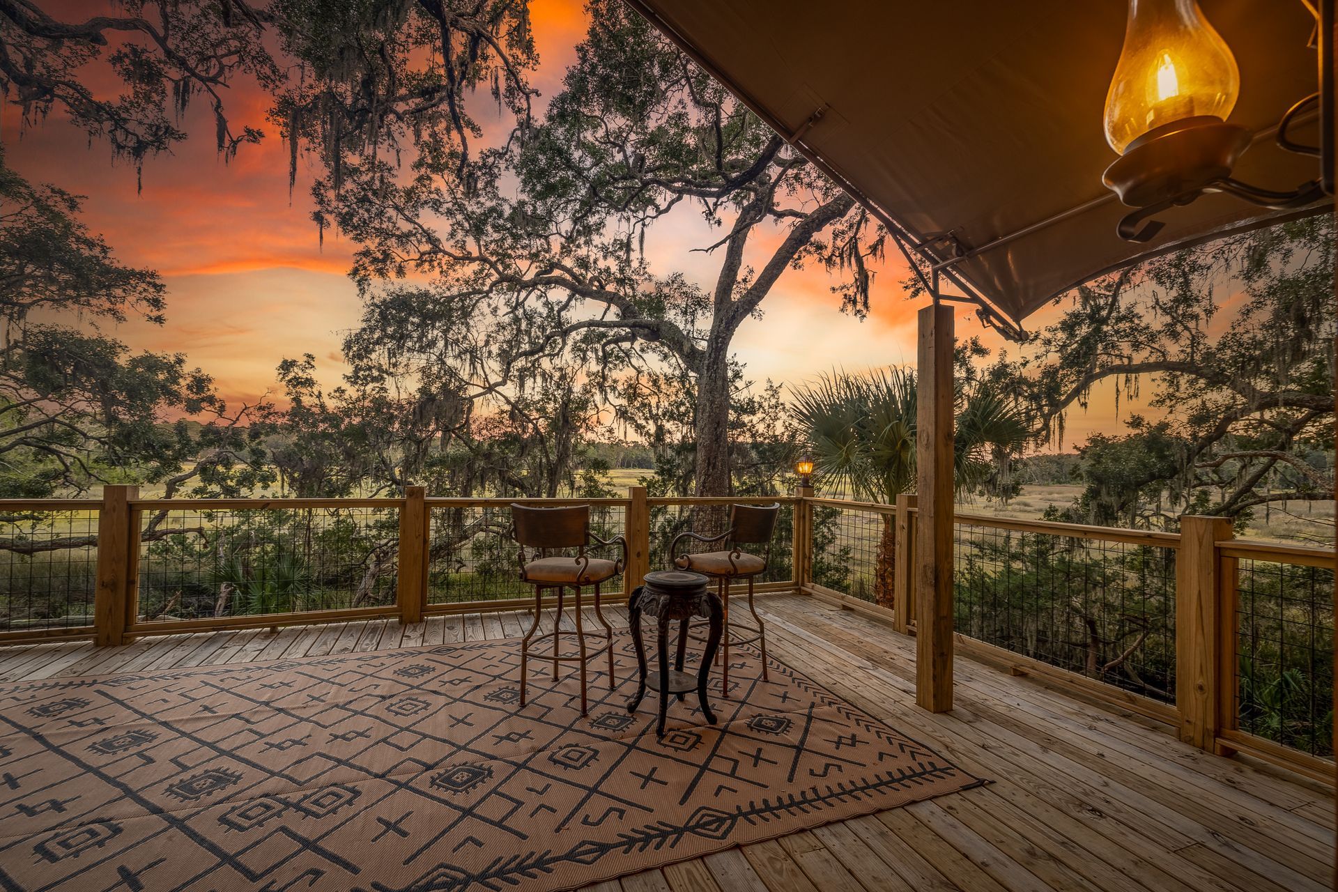 Wooden deck with chairs, rug, and lamp overlooking a field at sunset.