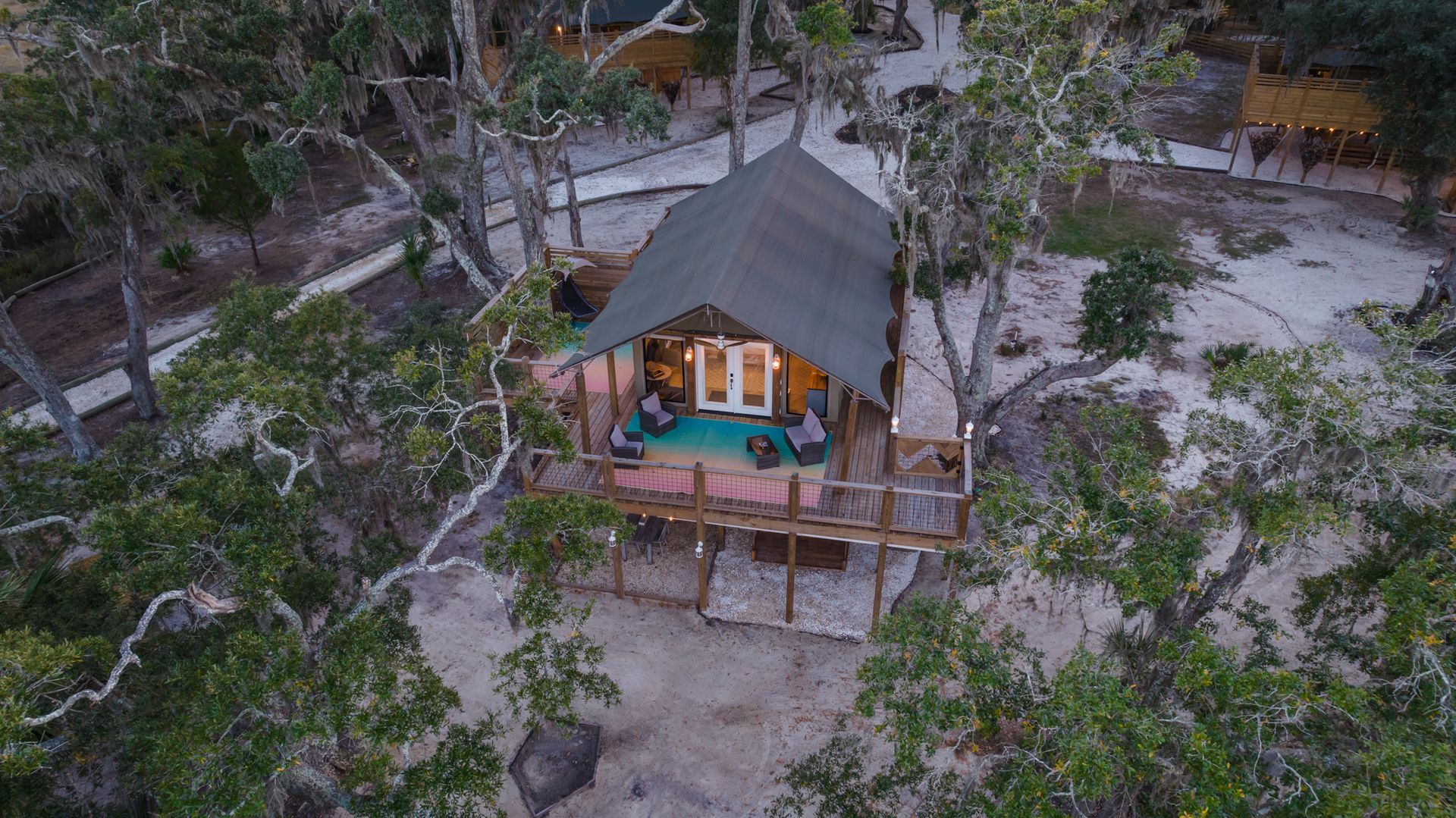 Aerial view of a treehouse nestled among trees; a deck with a seating area; other structures visible.