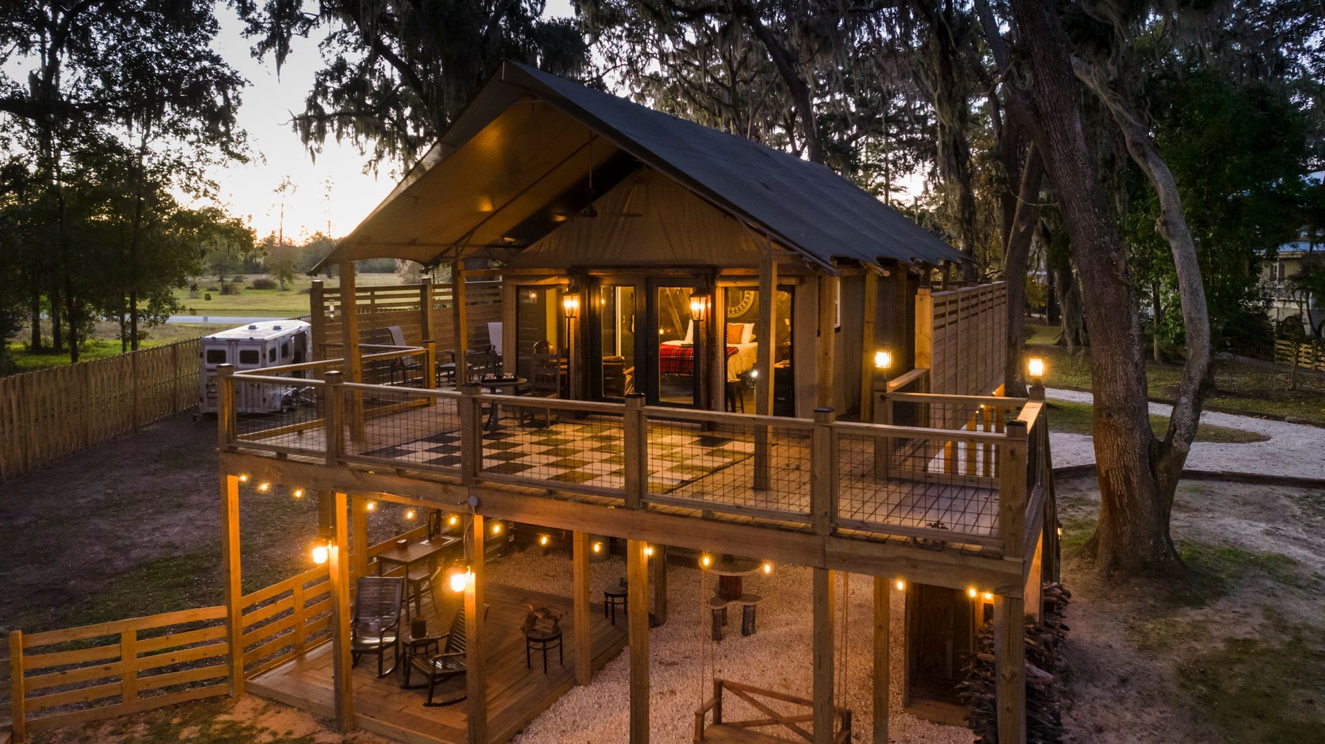 Wooden cabin with two-level deck lit with string lights at dusk. Trees surround.
