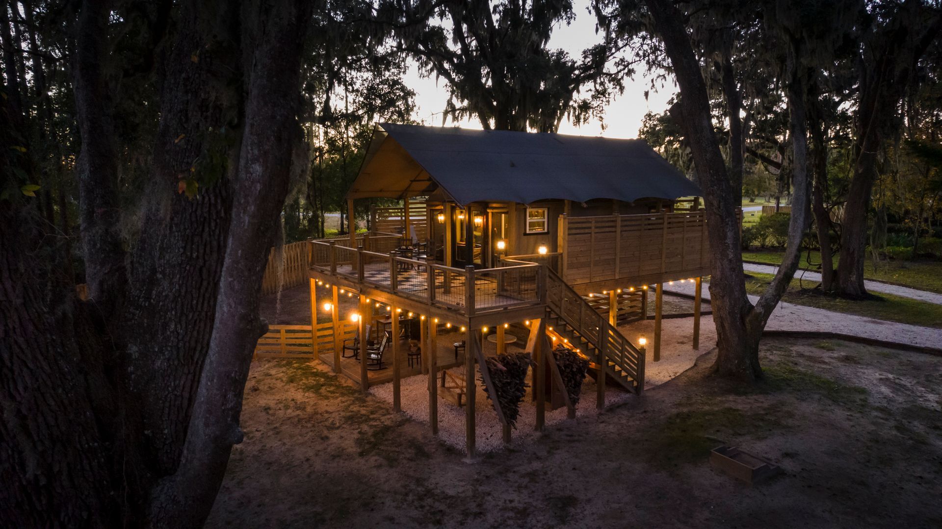 Treehouse at dusk, lit with string lights. Elevated wooden structure among trees.