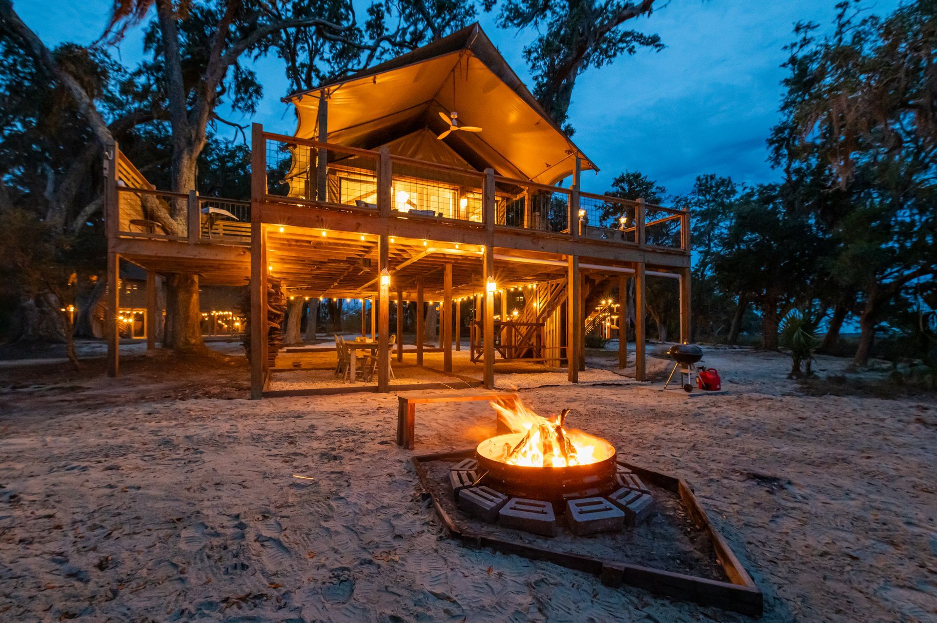 Lit treehouse with a deck, next to a campfire on a sandy ground at dusk.