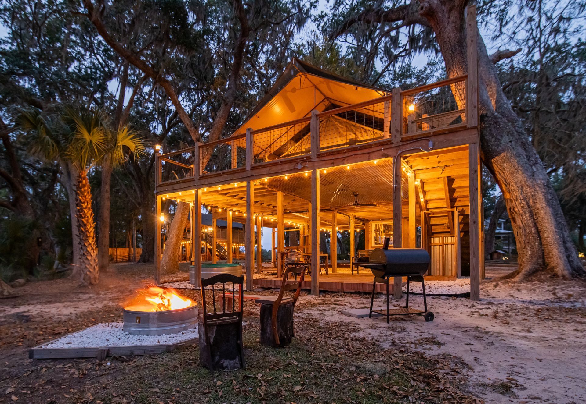 Lit wooden treehouse with string lights, fire pit, and grill in a wooded area at dusk.