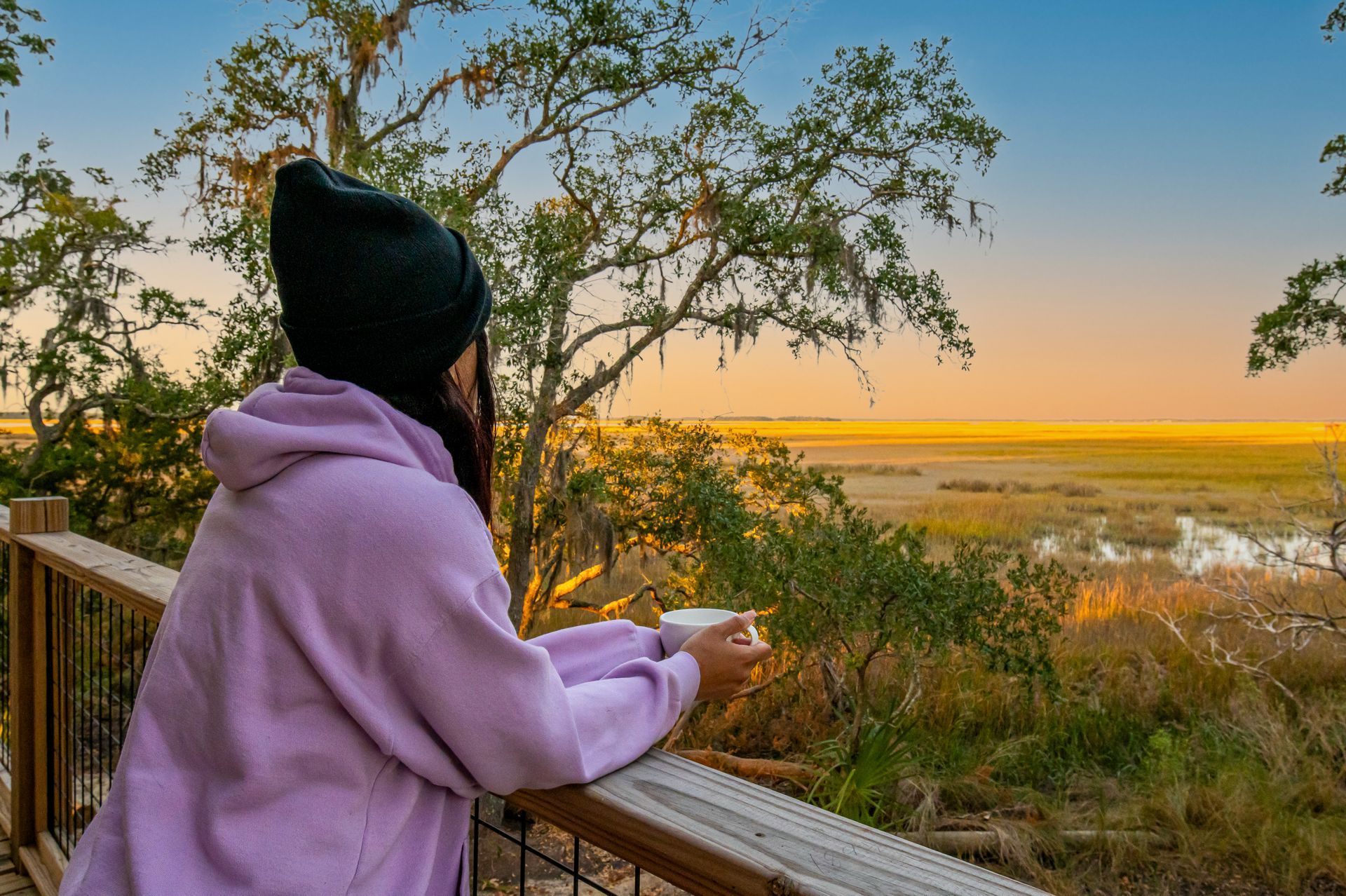 Person with a black hat and lavender hoodie, holding a mug, looks over a marsh at dawn.