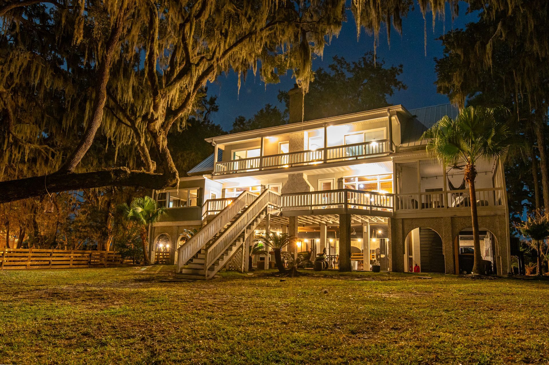 Lit-up two-story house with balconies at night, surrounded by trees and grass.