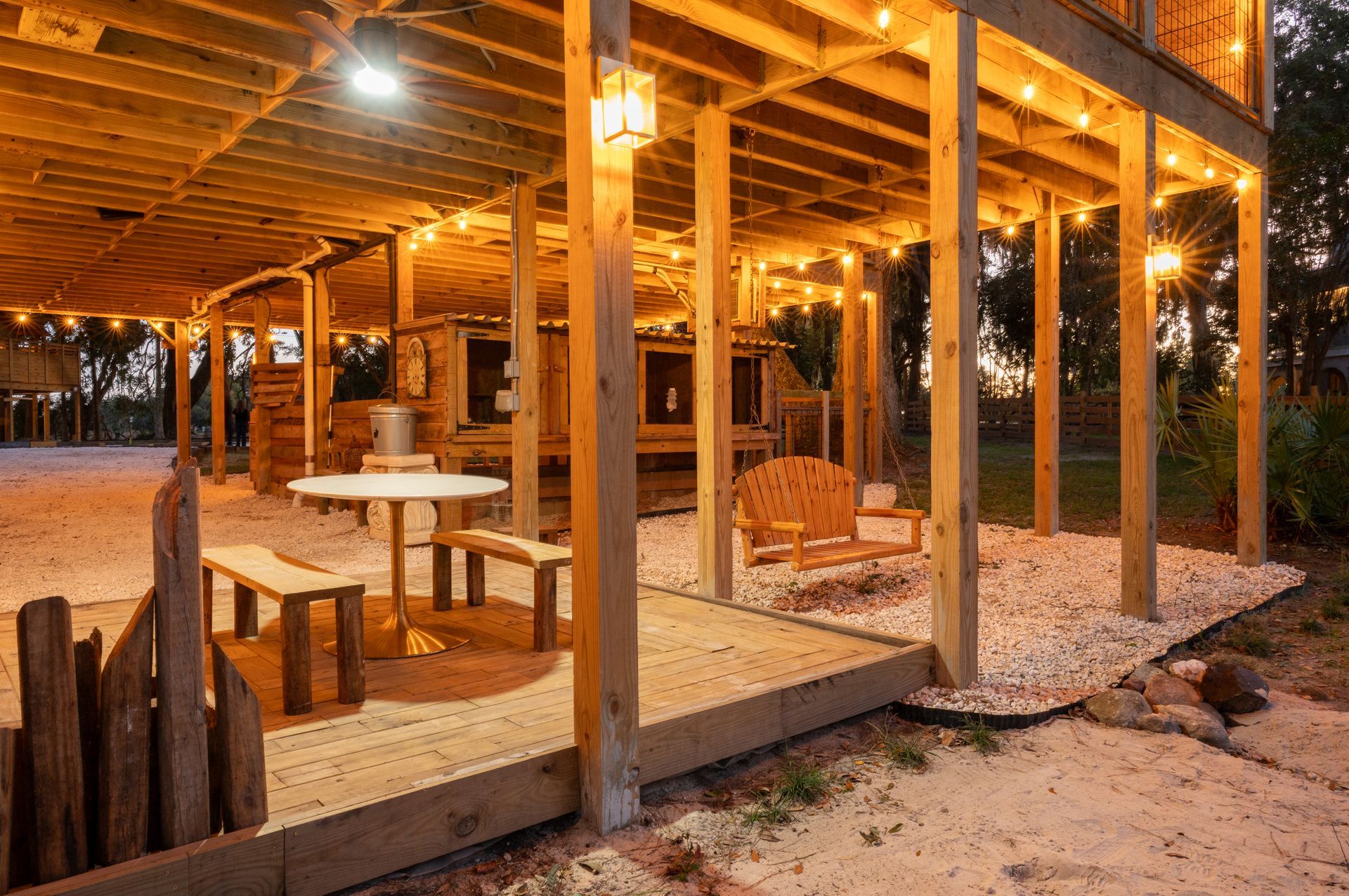 Outdoor wooden deck with string lights, table, benches, and a swing, all lit by evening light.