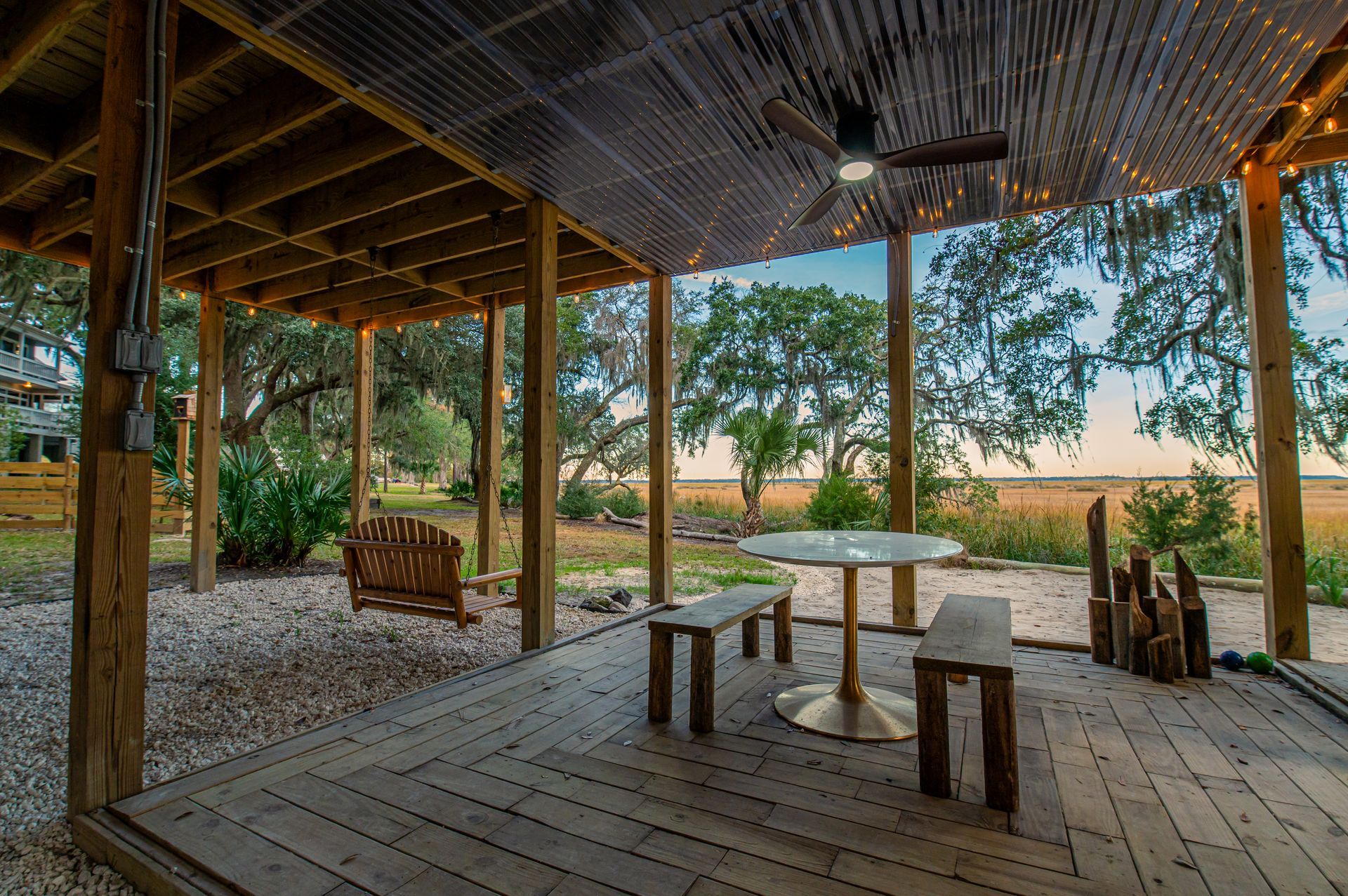 Patio with wooden beams, tables, benches, and a swing, overlooking a field and trees.