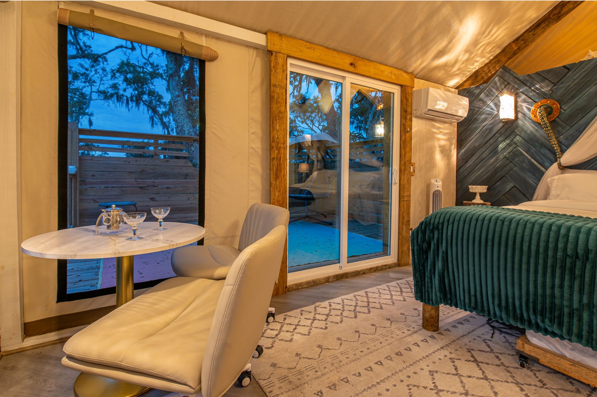Glamping tent interior with sliding glass door, table, chair, bed, and decorative wall.