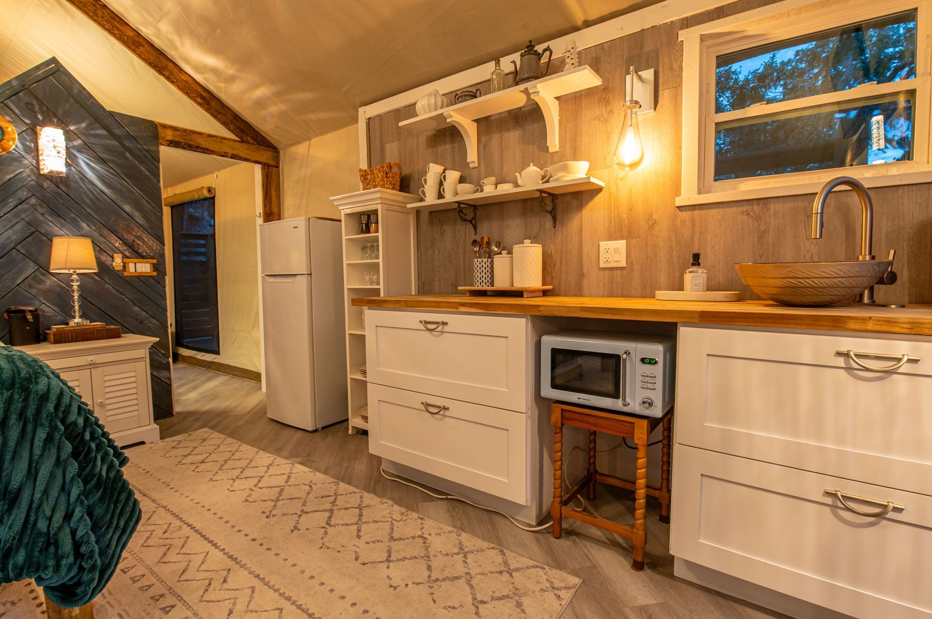Small cabin interior with a kitchen area and bed; white cabinets, microwave, sink, and window.