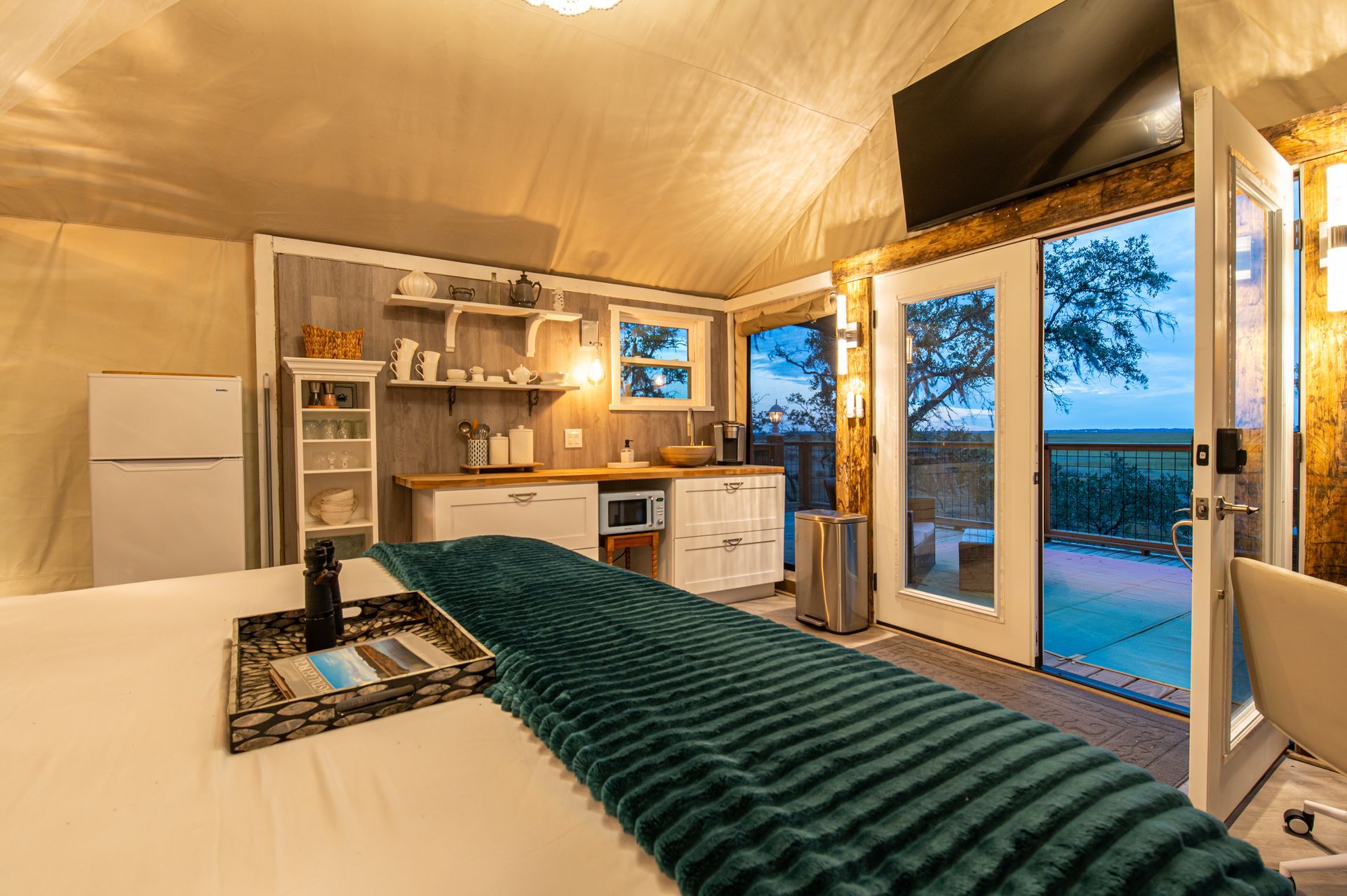Cozy glamping interior: bed, kitchenette, and open doorway to outdoor view. Teal throw, white fridge.