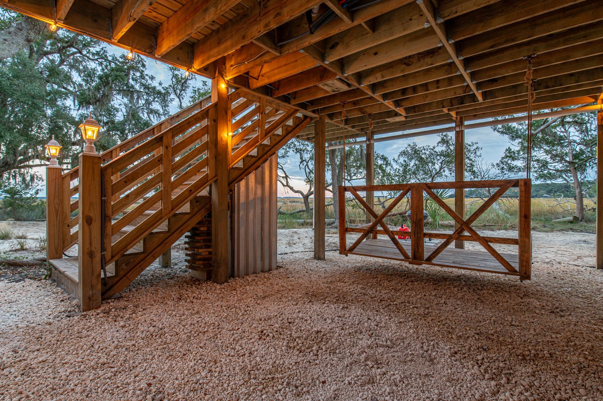 Wooden stairs under a raised deck lead to an outdoor area with gravel flooring. Lanterns illuminate the stairs.