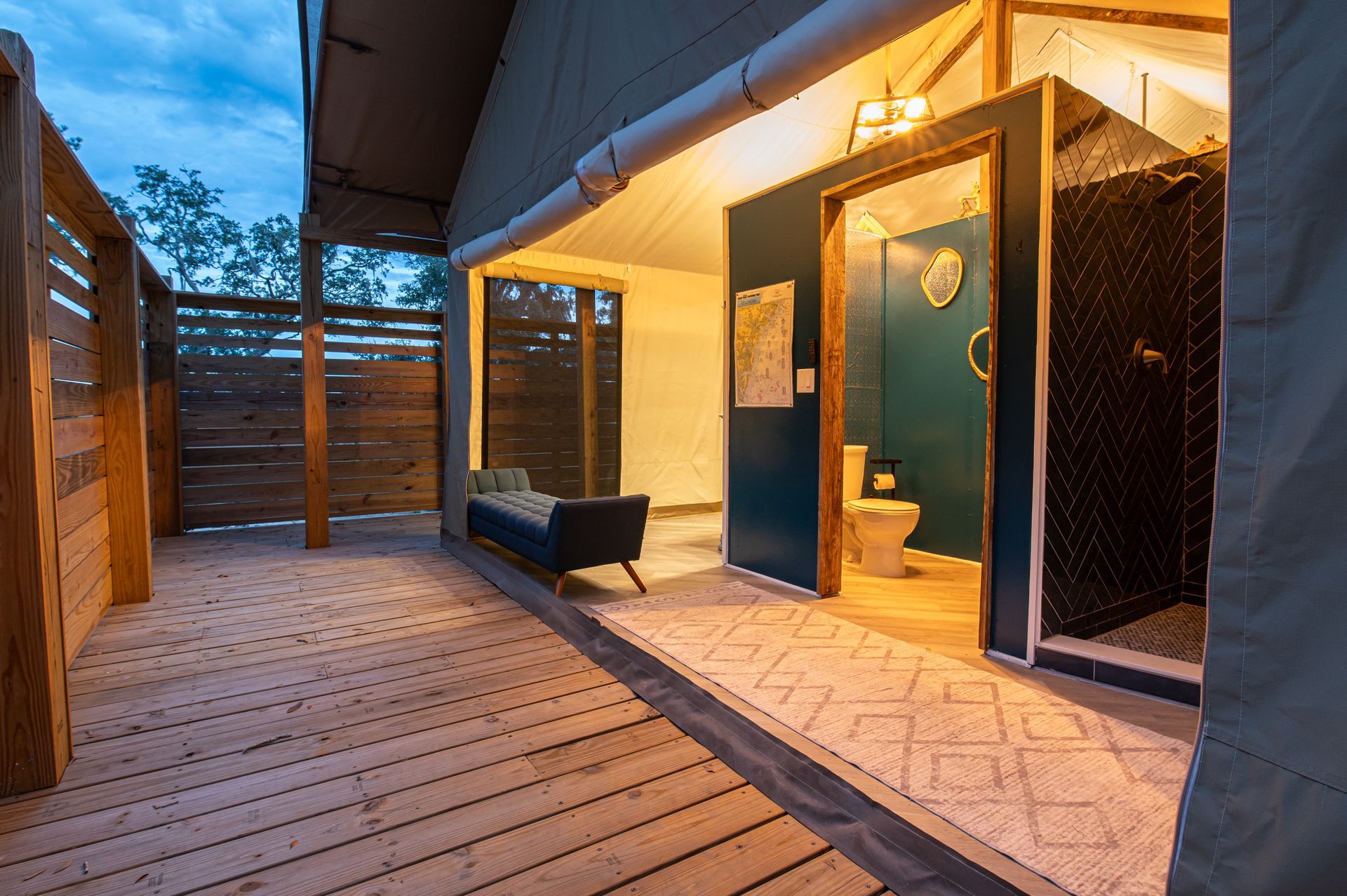 Wooden deck leading to open-air bathroom with turquoise door, bench, and shower, under a canvas tent roof.