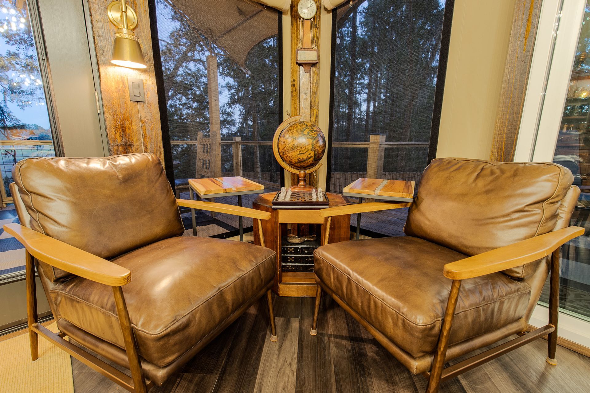 Two leather armchairs face each other near a table with a globe, inside a room with large windows and trees visible.