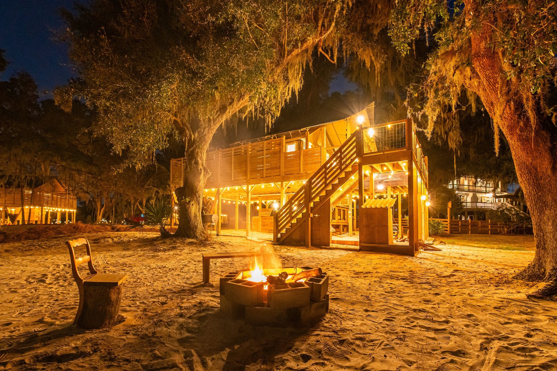 Campground at dusk with bonfire, wooden structures, and trees with lights.