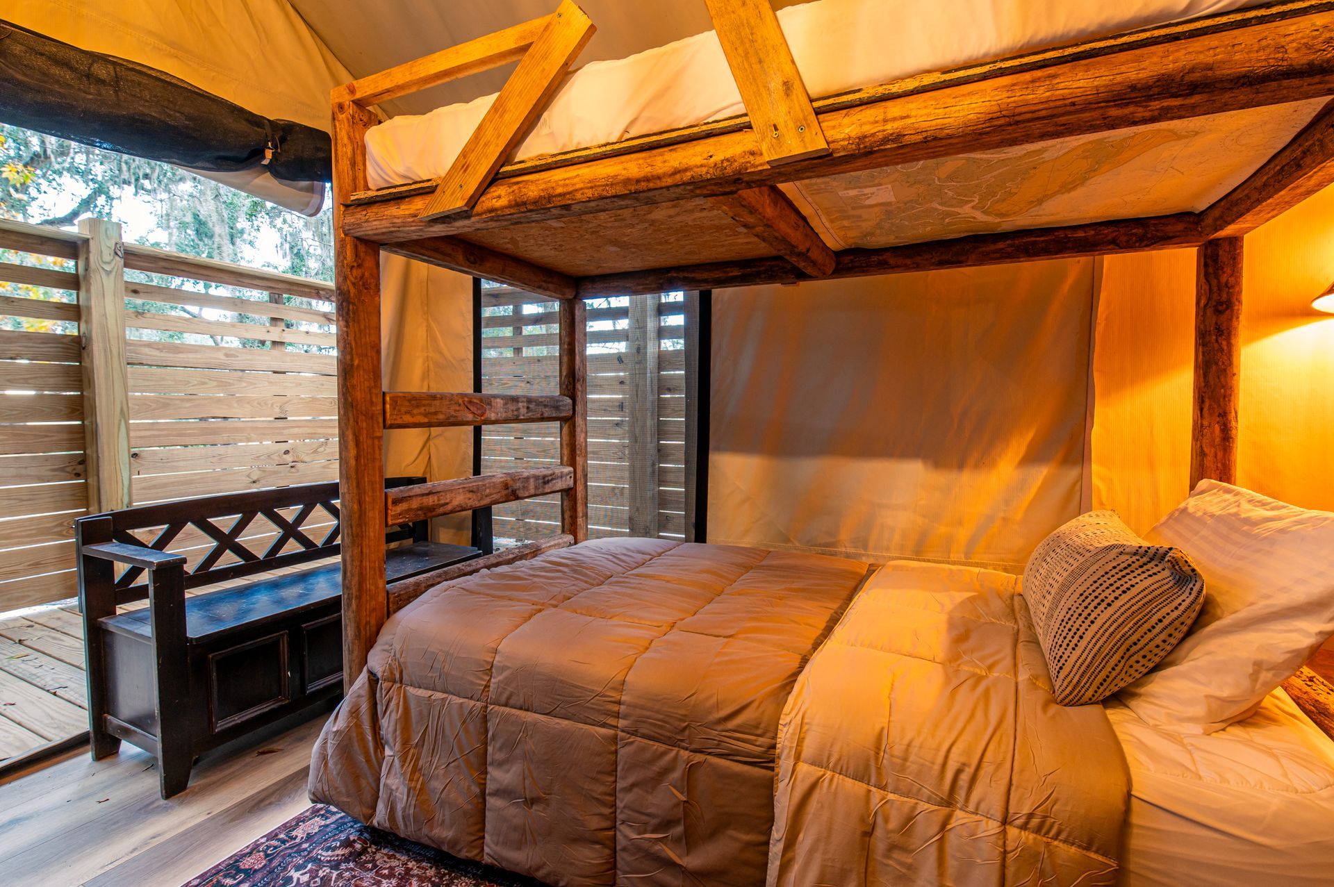 A tent interior with a bed and a bunk bed. Warm lighting, a bench, and a view of the outside.
