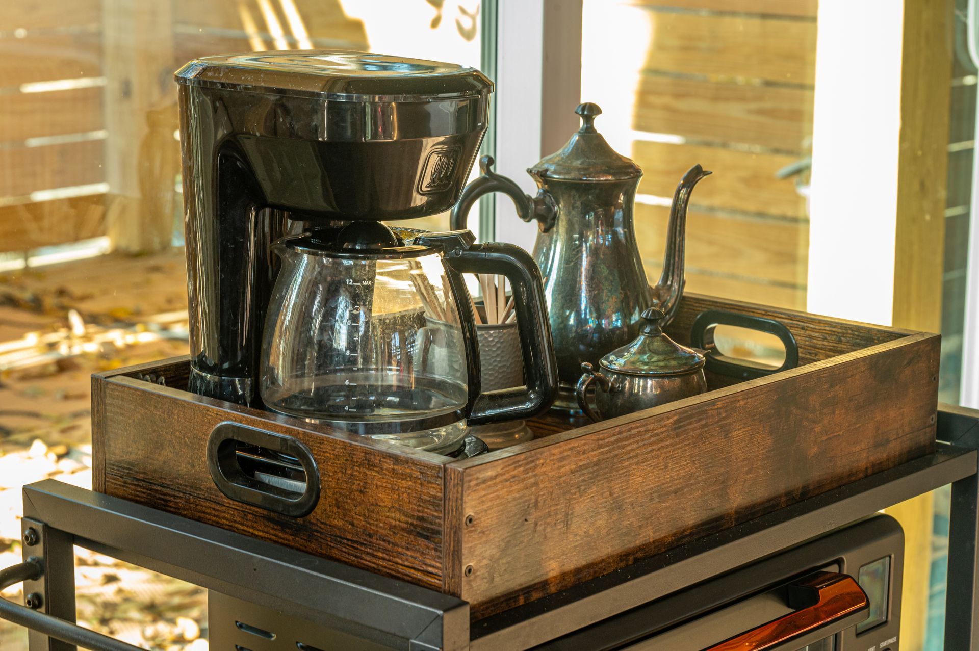 Coffee maker and antique silver server set on a tray, next to a window.