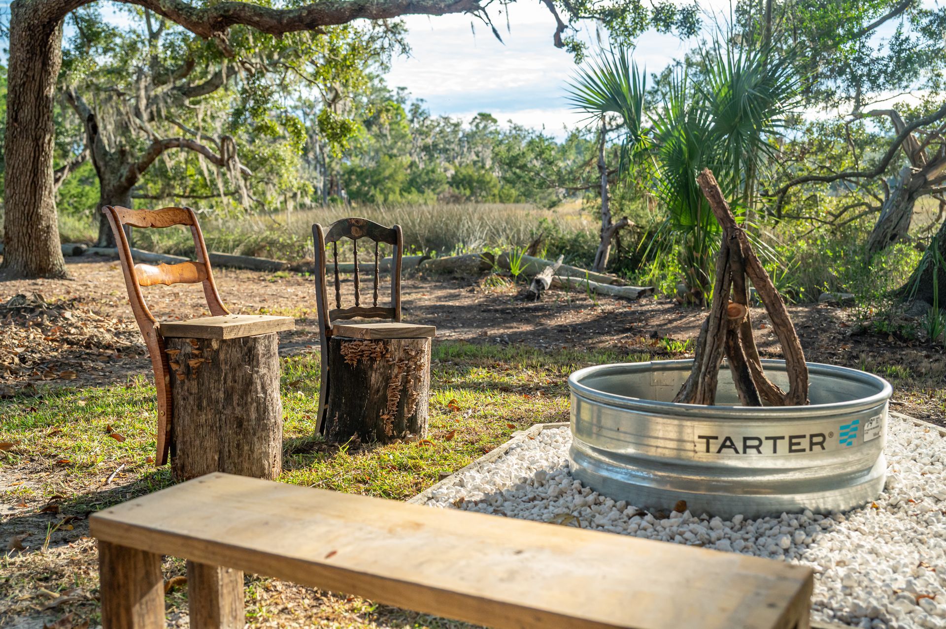 Outdoor fire pit with chairs and bench in a natural setting.
