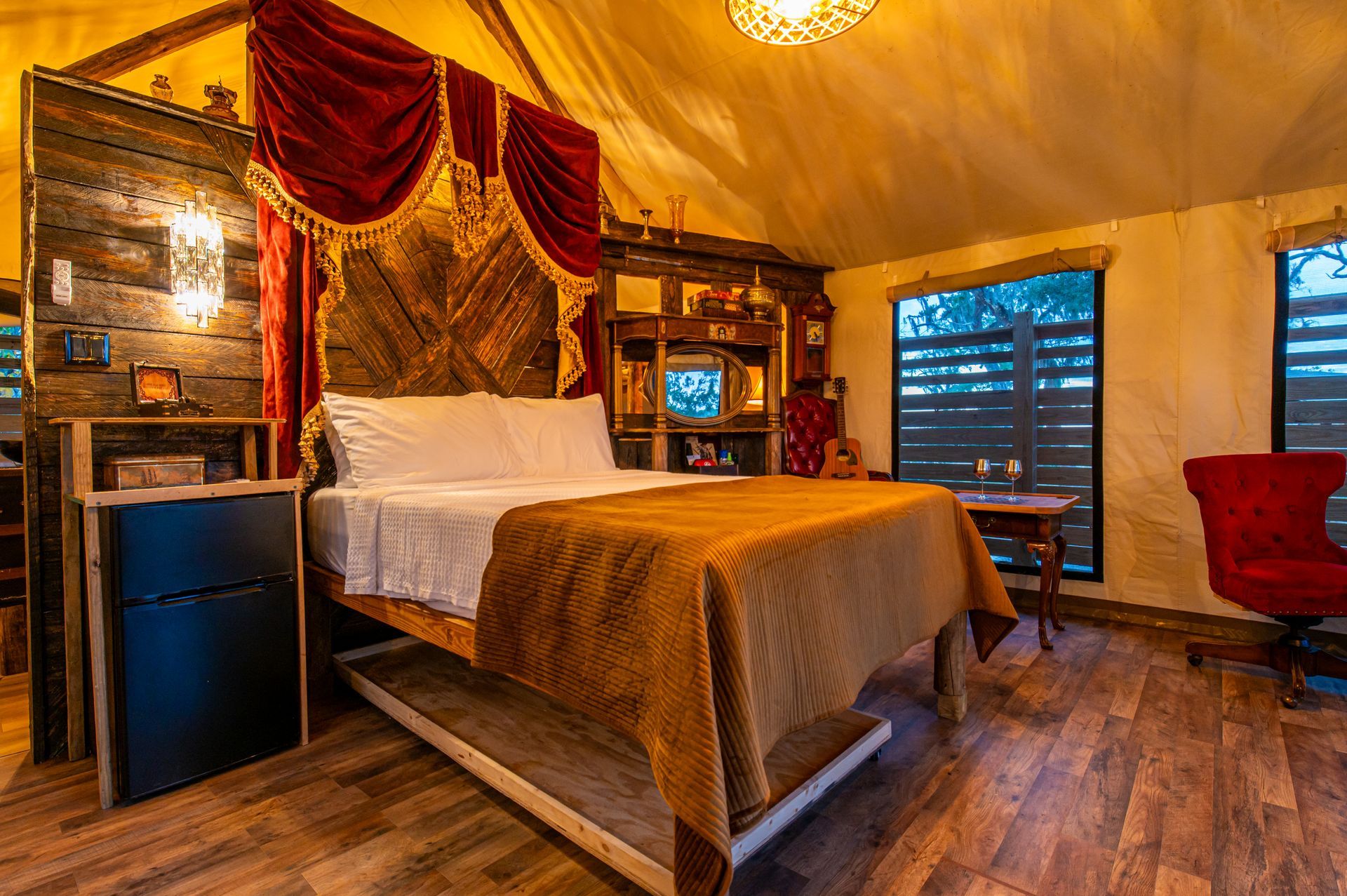 Cozy tent interior with bed, bookshelves, and red velvet curtain. Window view. Warm lighting and wood flooring.