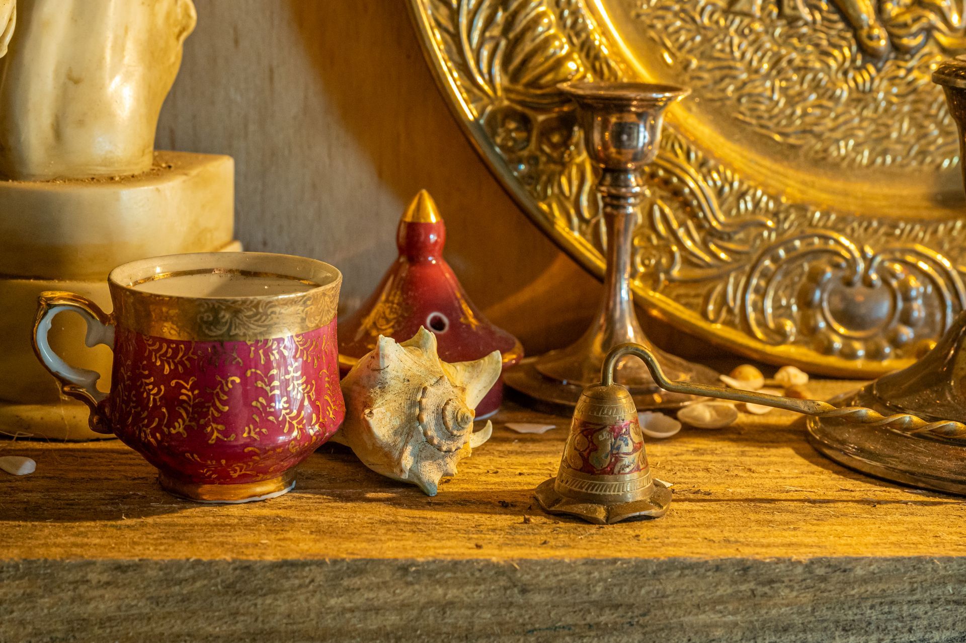 Red and gold cup, seashell, candlestick, and ornate gold plate on a wooden shelf, lit by warm light.