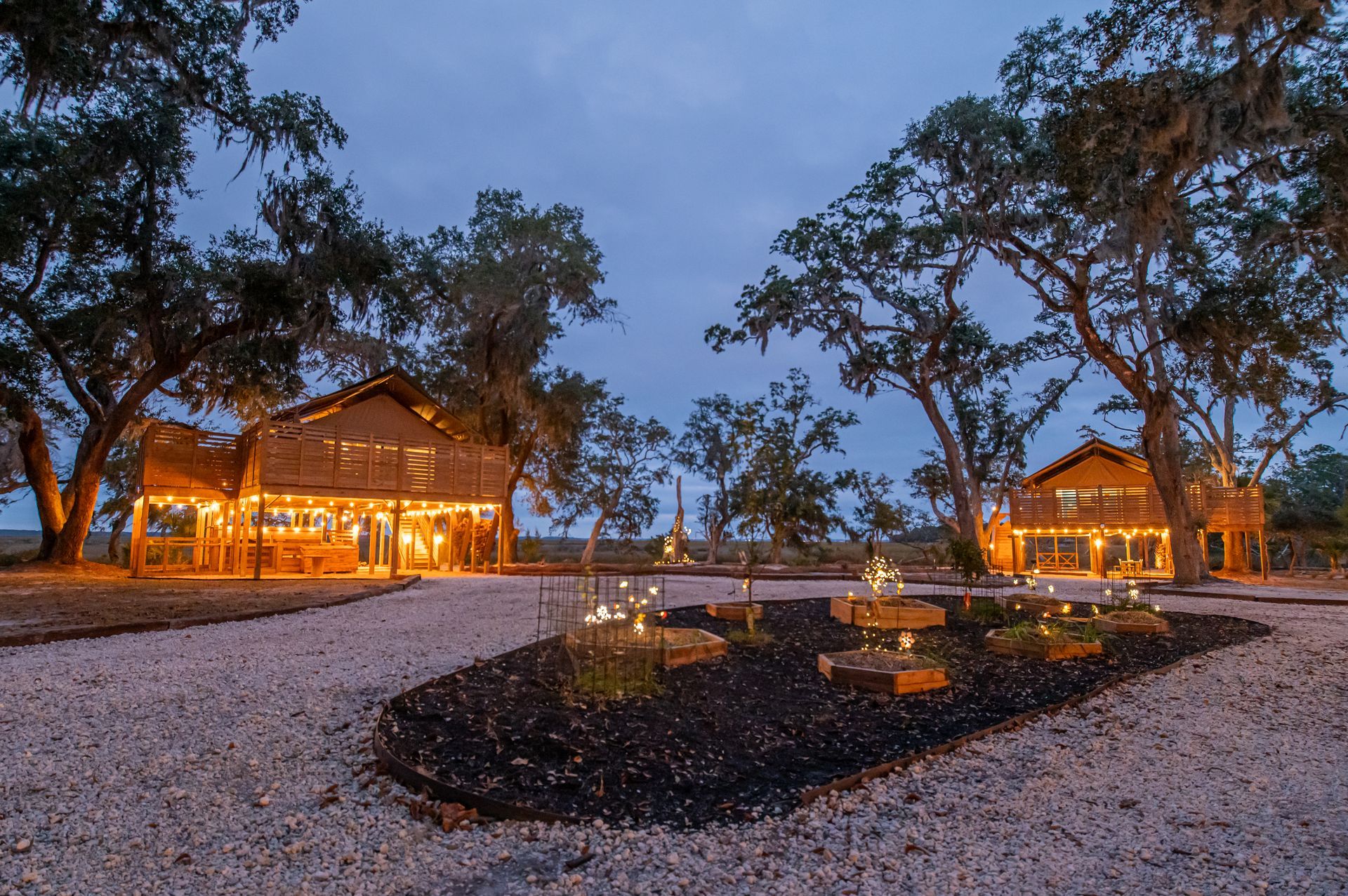 Two wood-framed buildings at dusk with string lights and trees, set in a gravel area with a dark flower bed.