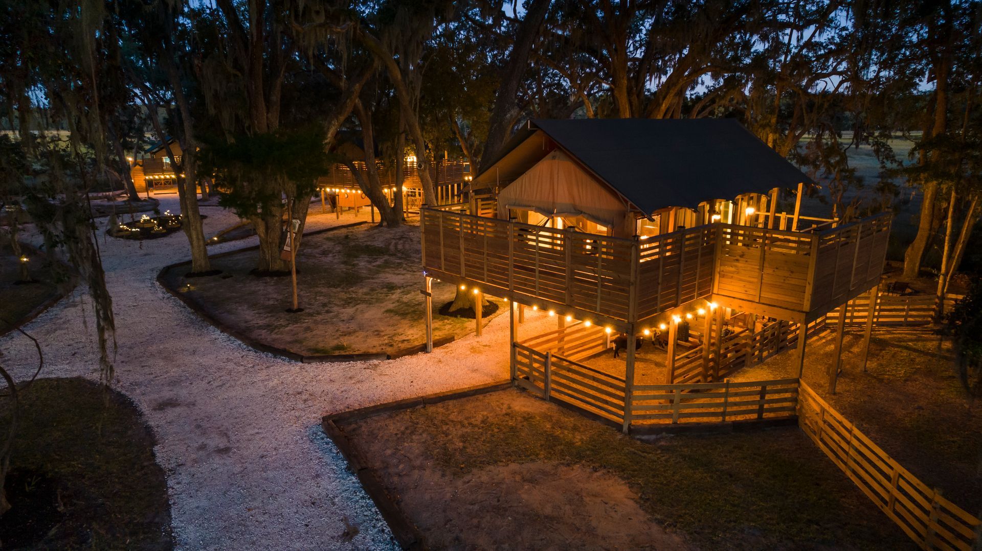 Lit treehouse at dusk with string lights, wooden deck, and gravel path.