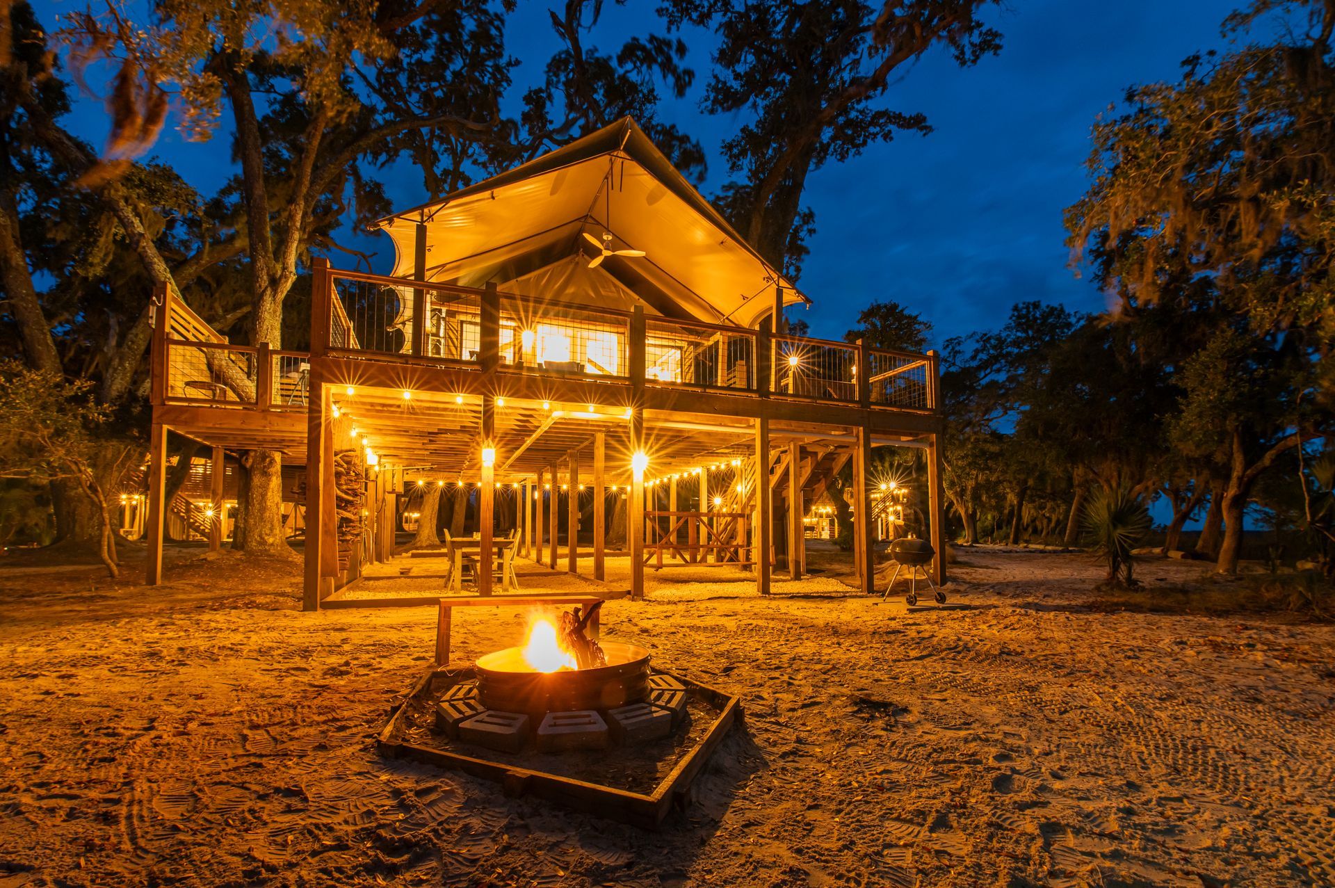 Wooden treehouse lit with string lights at night, with a fire pit in the foreground.