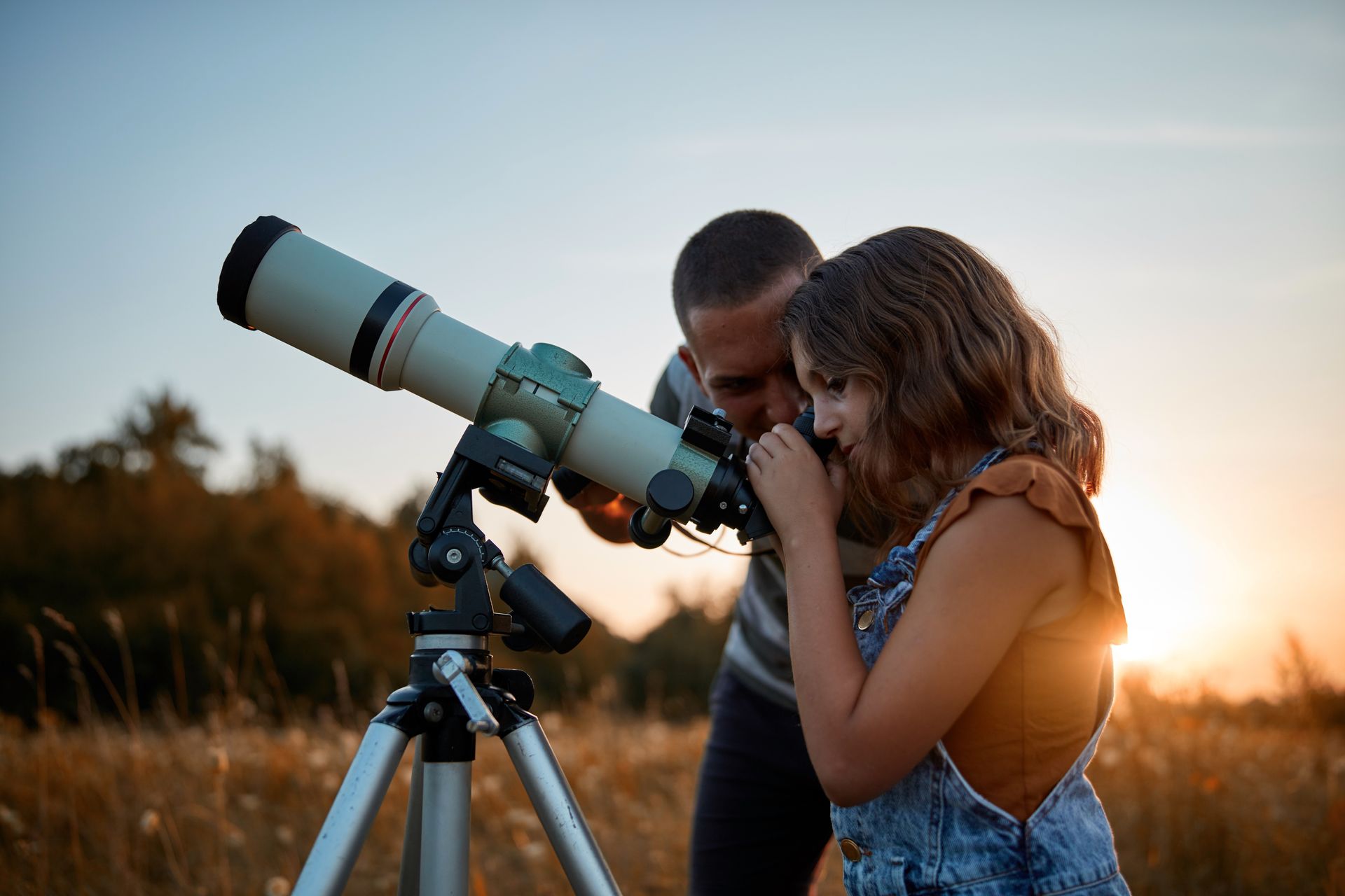 Man and girl looking through a telescope at sunset in a field of tall grass.