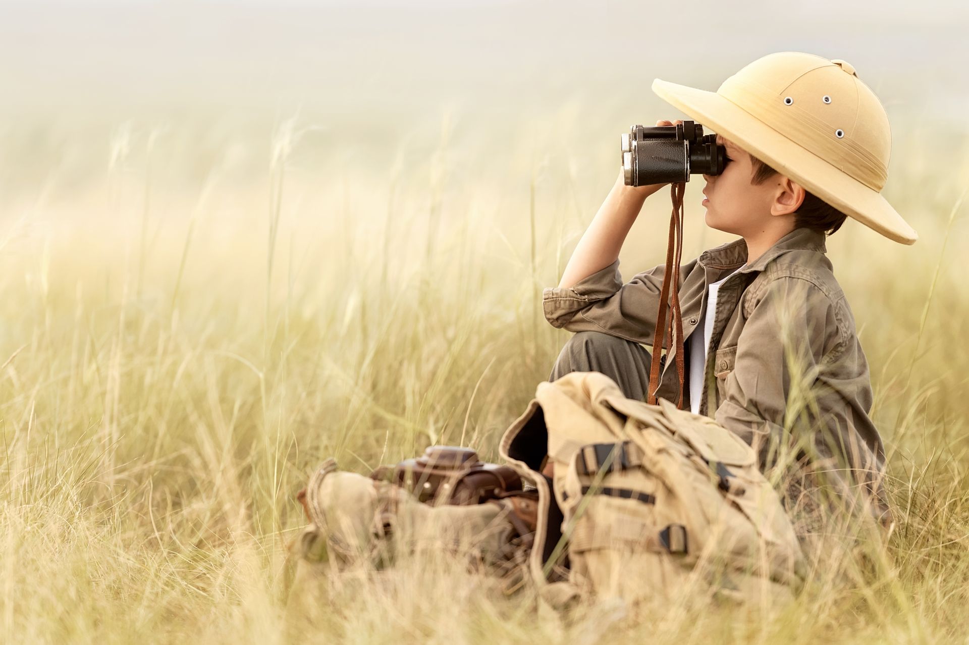 Boy with binoculars wearing a safari hat, looking through tall grass. Backpack beside him.