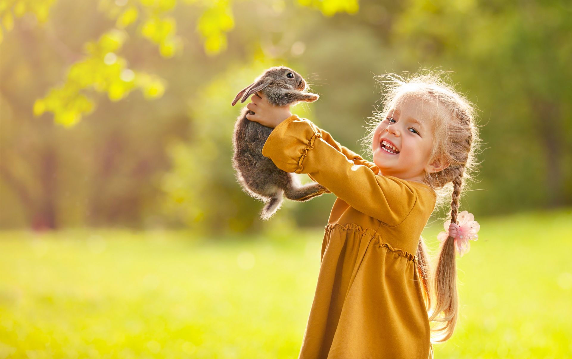 Girl in mustard dress joyfully holds a rabbit in a sunny green field.